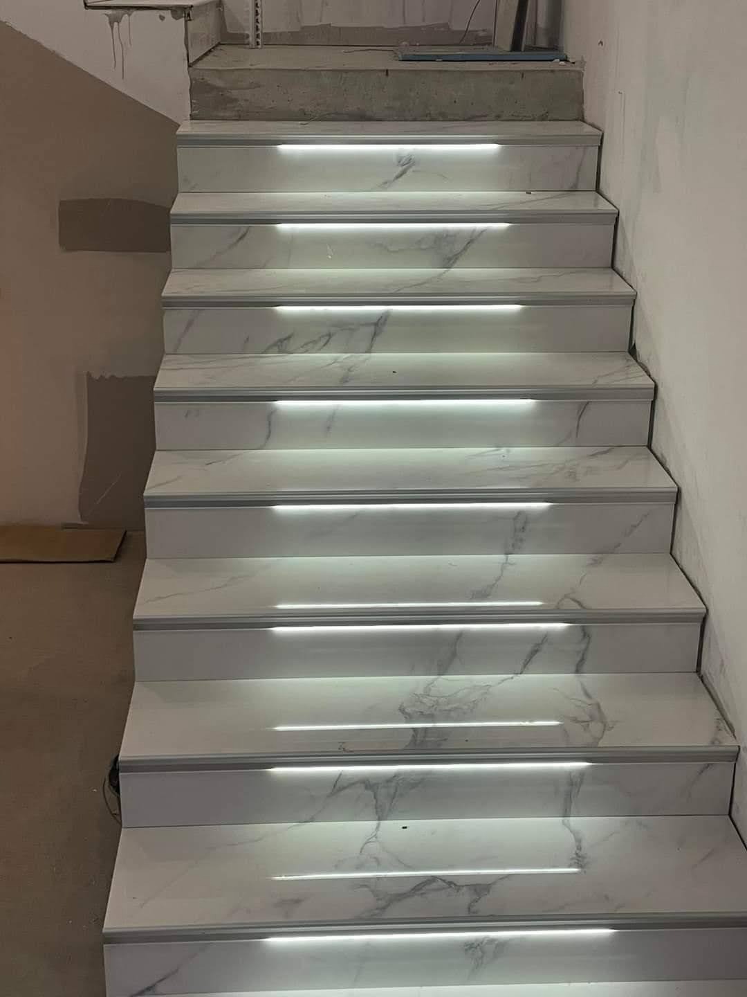 Schody wyłożone białym marmurem z subtelnym podświetleniem LED, tworzące nowoczesny i elegancki efekt wizualny. Widok z dołu, podkreślający detale wykończenia.