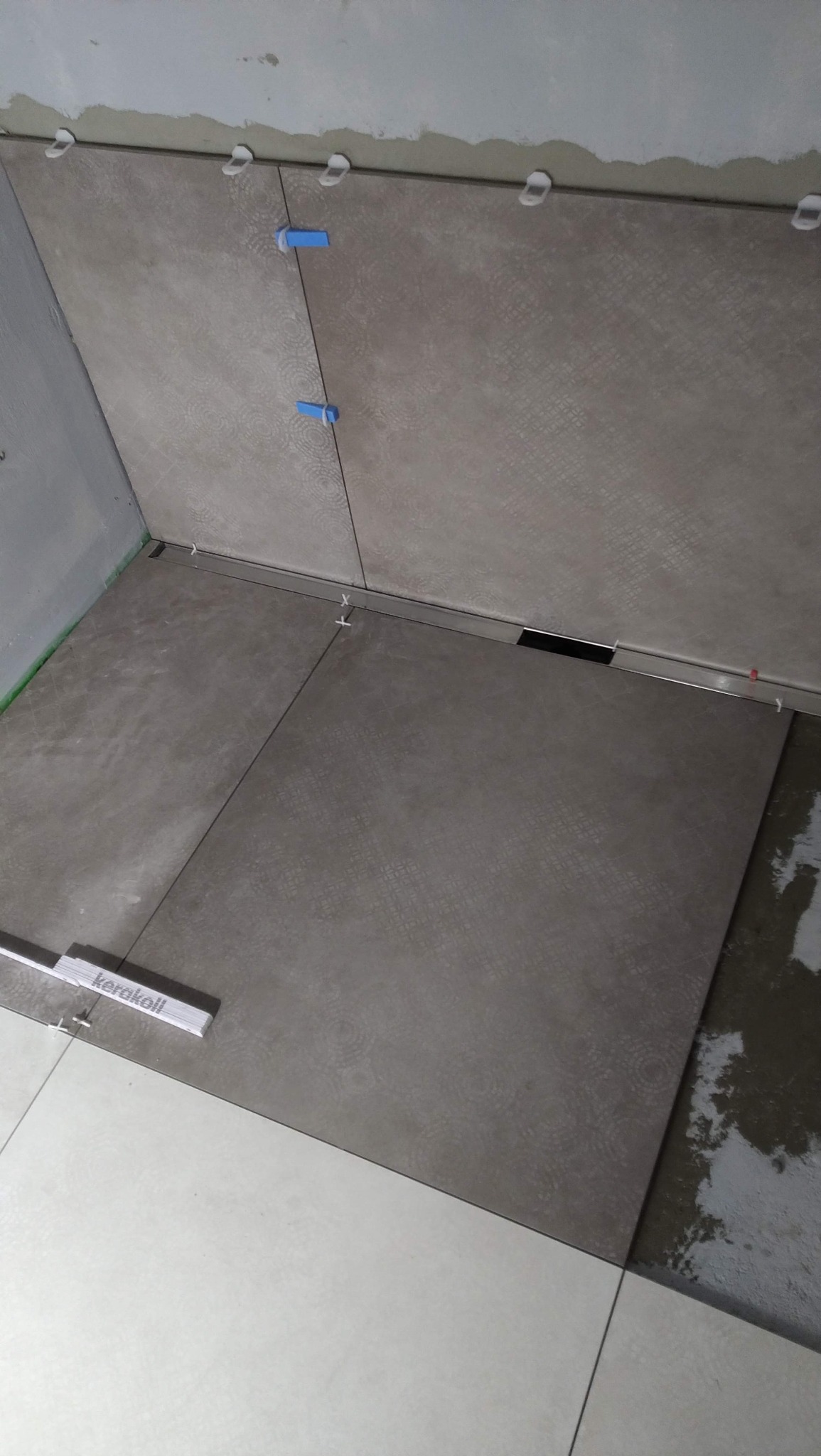 Układanie płytek podłogowych w łazience, widoczne narzędzia glazurnicze i fragment ułożonej podłogi. Szare płytki z delikatnym wzorem, białe fugi.