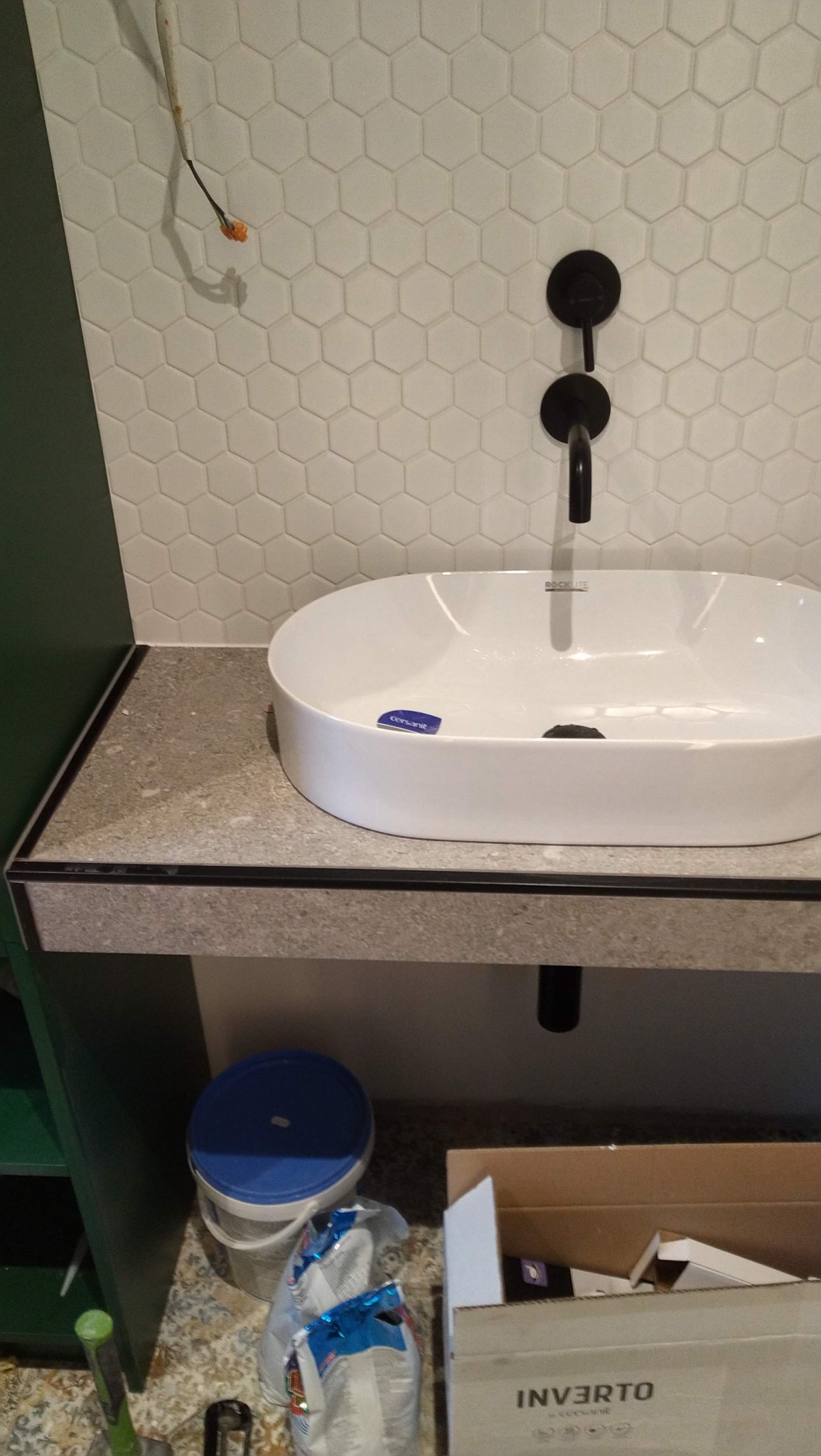 Biała umywalka nablatowa Cersanit na blacie z szarego kamienia, czarna bateria, heksagonalne płytki na ścianie. Widoczne materiały budowlane pod blatem.