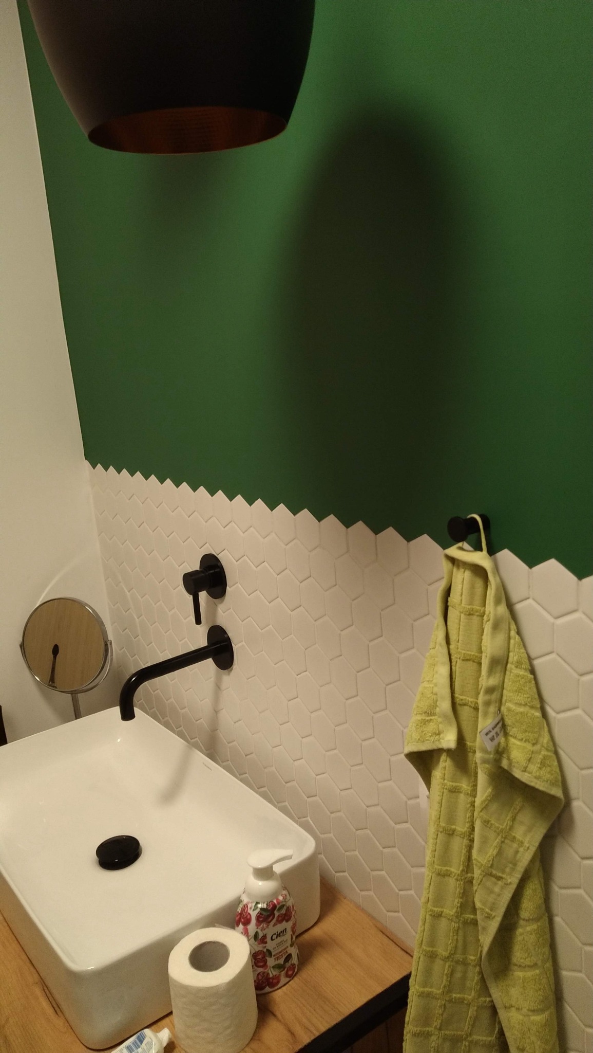 Nowoczesna łazienka z zieloną ścianą, białymi heksagonalnymi płytkami, czarną armaturą i drewnianym blatem pod umywalką. Żółty ręcznik dodaje koloru.