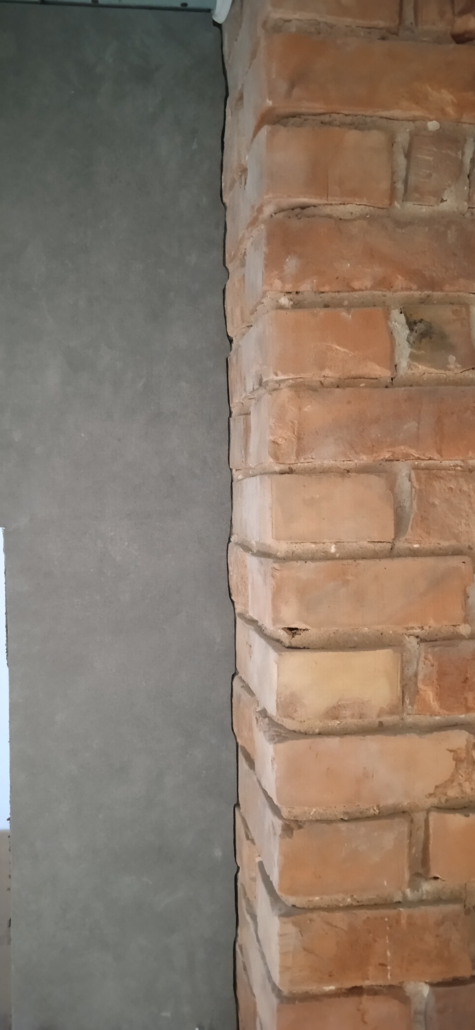 Fragment ściany z surowym tynkiem sąsiadującym z dekoracyjną ścianą z czerwonej cegły, ukazujący kontrast faktur i materiałów wykończeniowych.