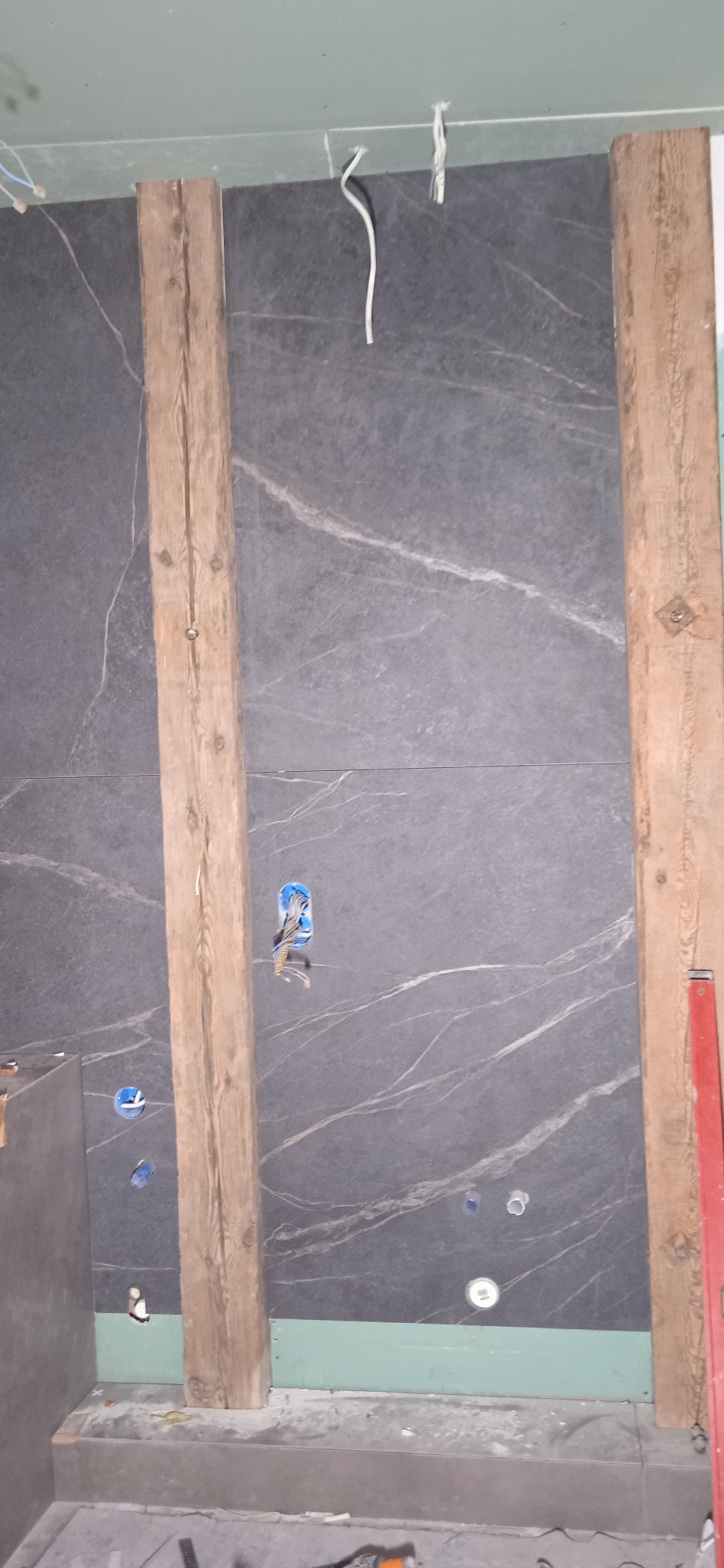 Ciemnoszara ściana z marmurowym wzorem w trakcie montażu płytek, z widocznymi drewnianymi elementami konstrukcyjnymi i instalacją elektryczną w otworach.