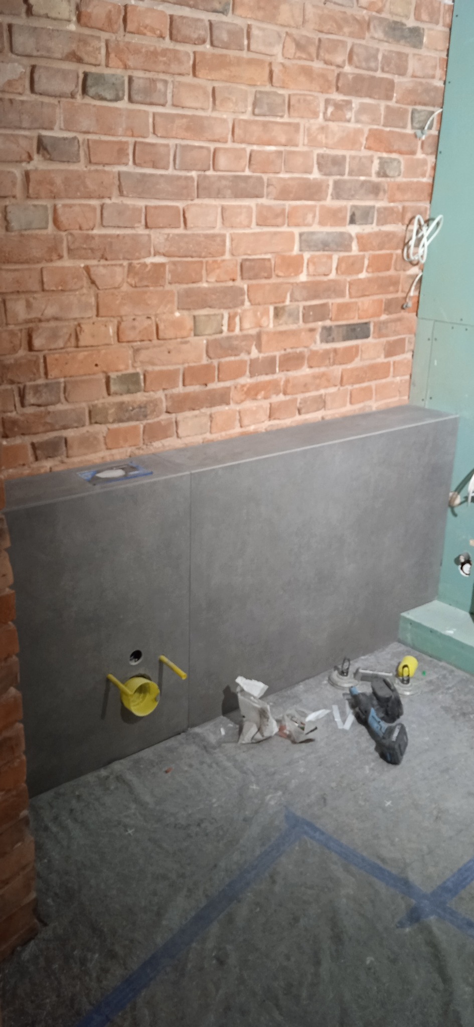 Ściana z cegły i szarej płyty w łazience w trakcie prac. Widoczne narzędzia i elementy instalacji. Surowy, industrialny styl.