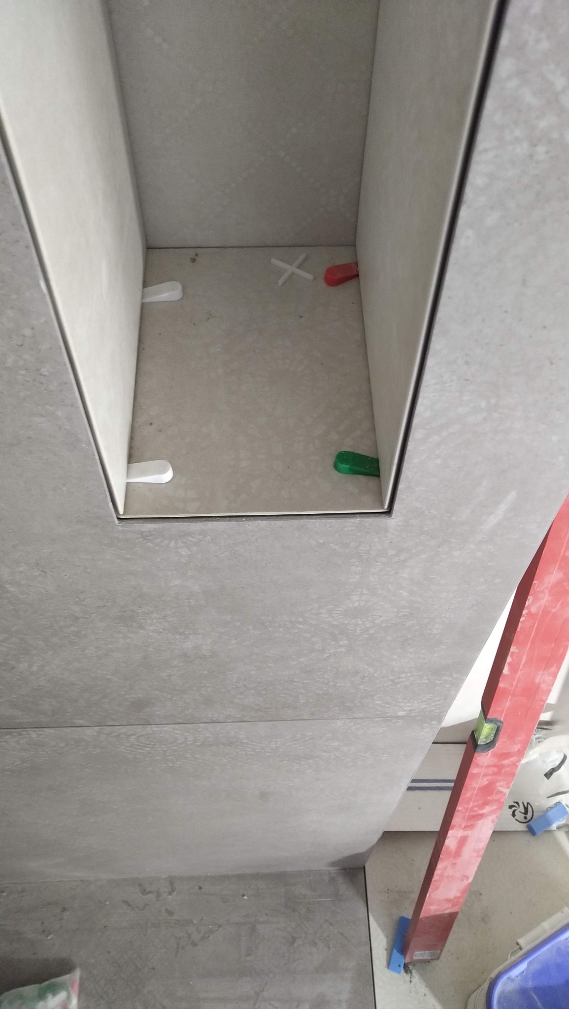 Wnęka wyłożona jasnymi, matowymi płytkami z widocznymi krzyżykami dystansowymi i poziomnicą, ukazująca etap prac glazurniczych.