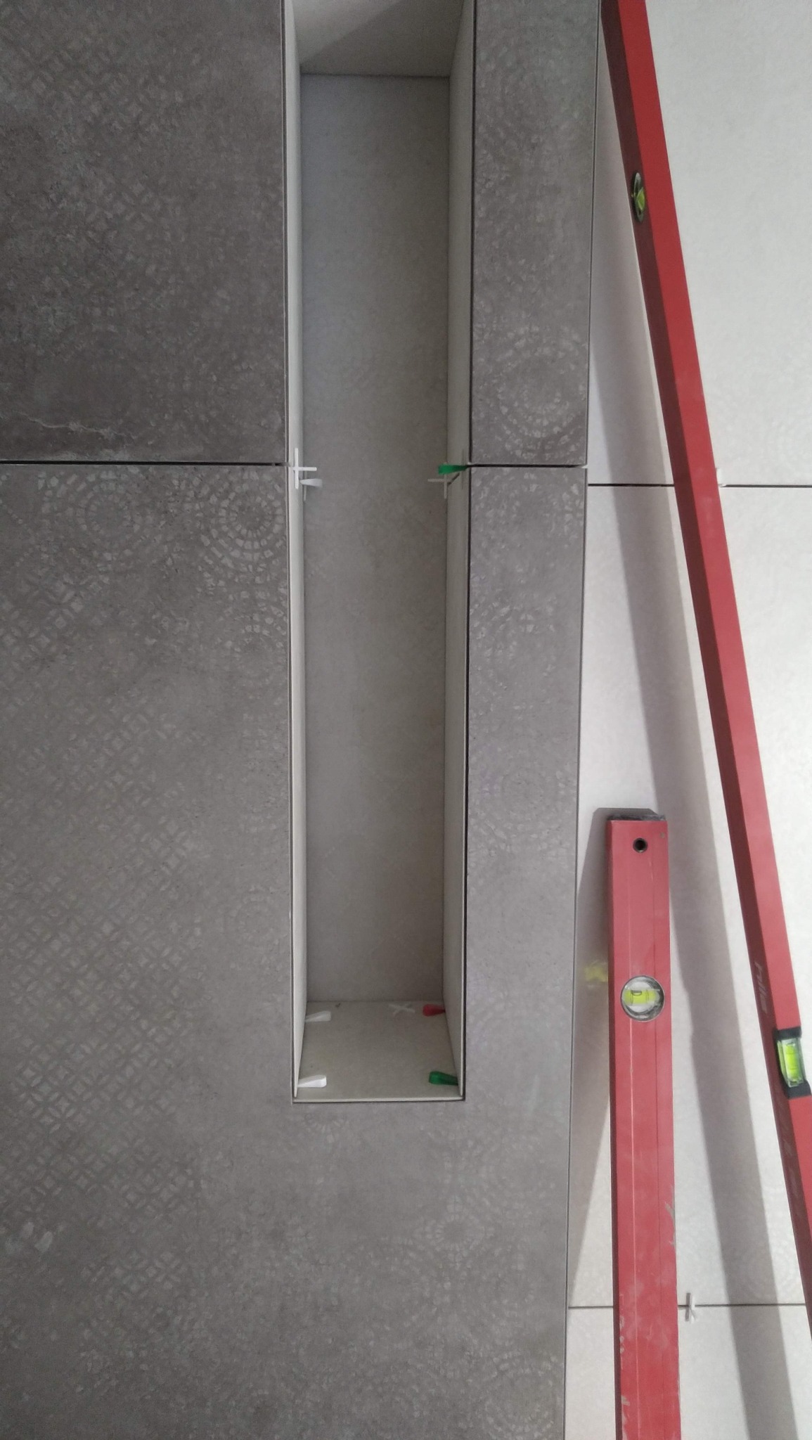 Szara ściana z płytek z wnęką, poziomica pionowo po prawej. Widoczne krzyżyki dystansowe. Wnęka wykończona płytkami.