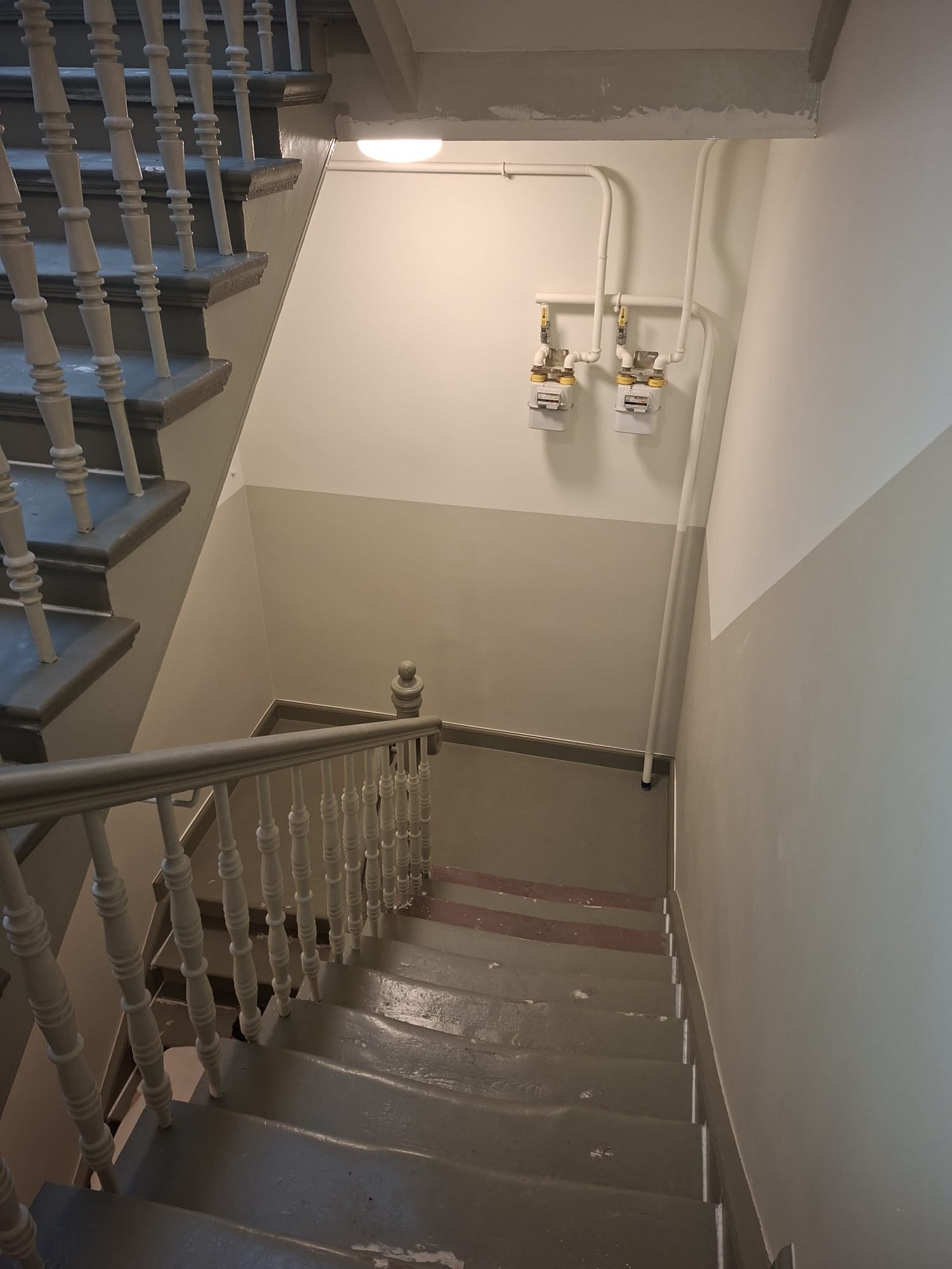 Klatka schodowa w kamienicy po odświeżeniu, widok z góry. Szare schody z drewnianą balustradą, pomalowane ściany z widocznymi licznikami gazu.