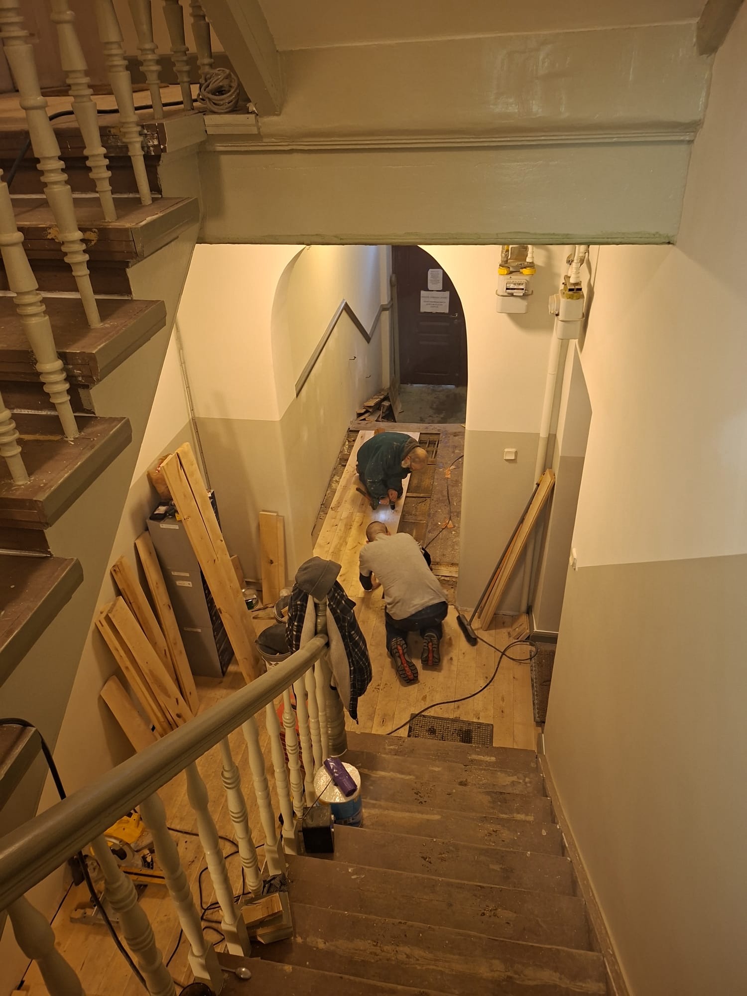 Dwóch mężczyzn montuje drewnianą podłogę na klatce schodowej w kamienicy. Widok z góry na etap prac. Widoczne narzędzia i materiały budowlane.