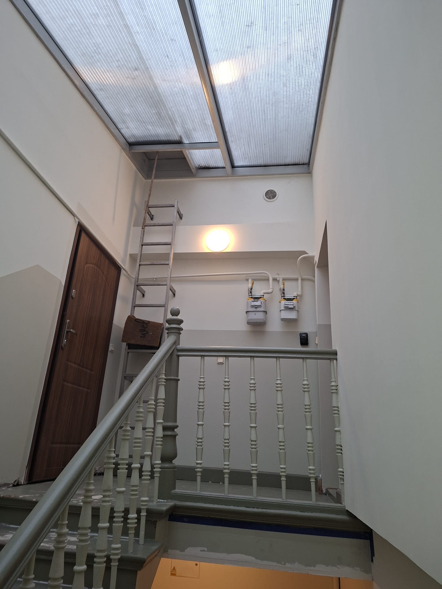 Klatka schodowa w trakcie prac wykończeniowych: drewniana balustrada, drabina oparta o ścianę, widoczne liczniki gazu i przeszklony sufit.