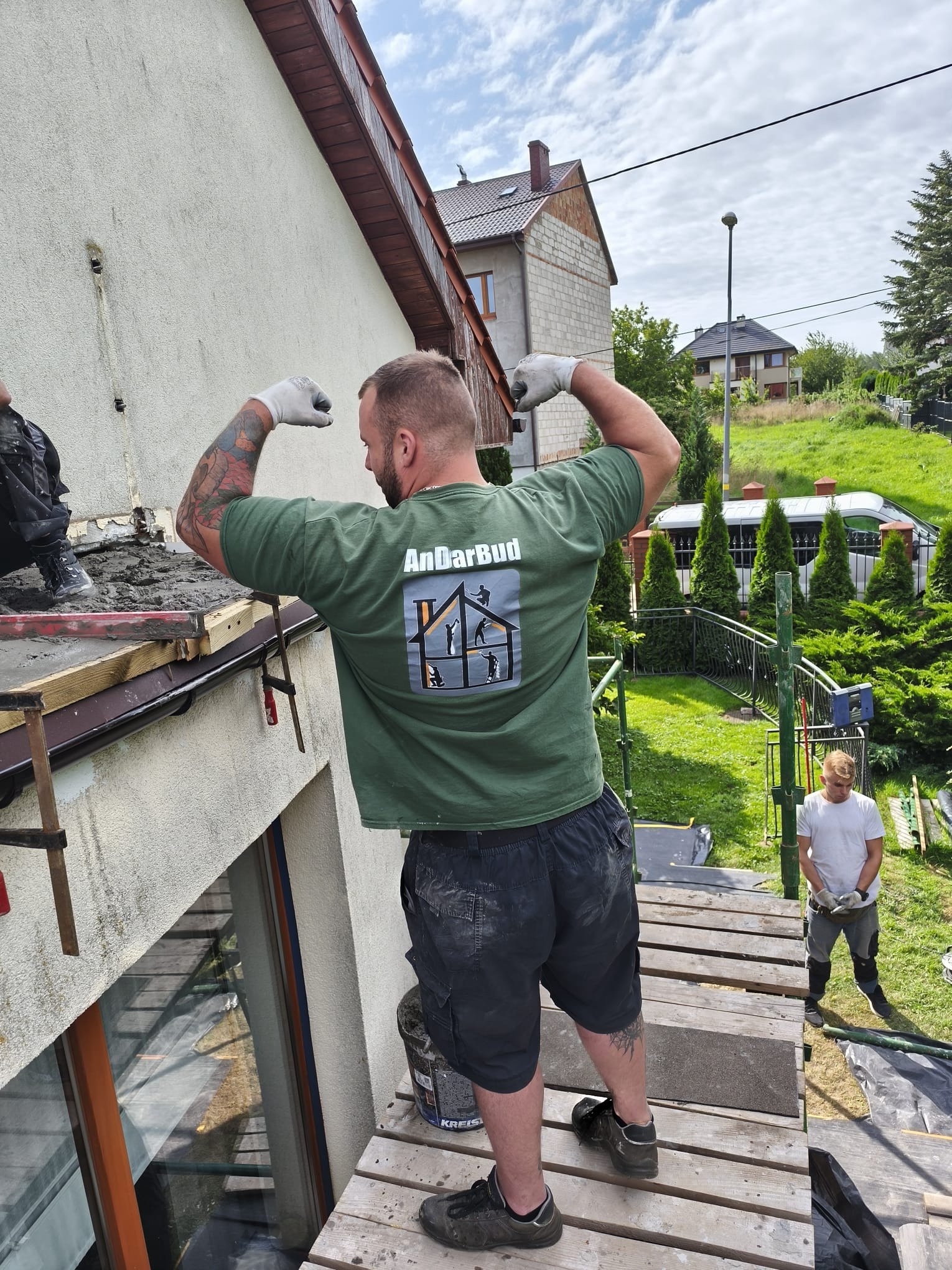 Pracownik w zielonej koszulce z logo firmy AndarBud prezentuje biceps na tle ocieplanej ściany domu. Widoczne rusztowanie, narzędzia i drugi pracownik w tle.