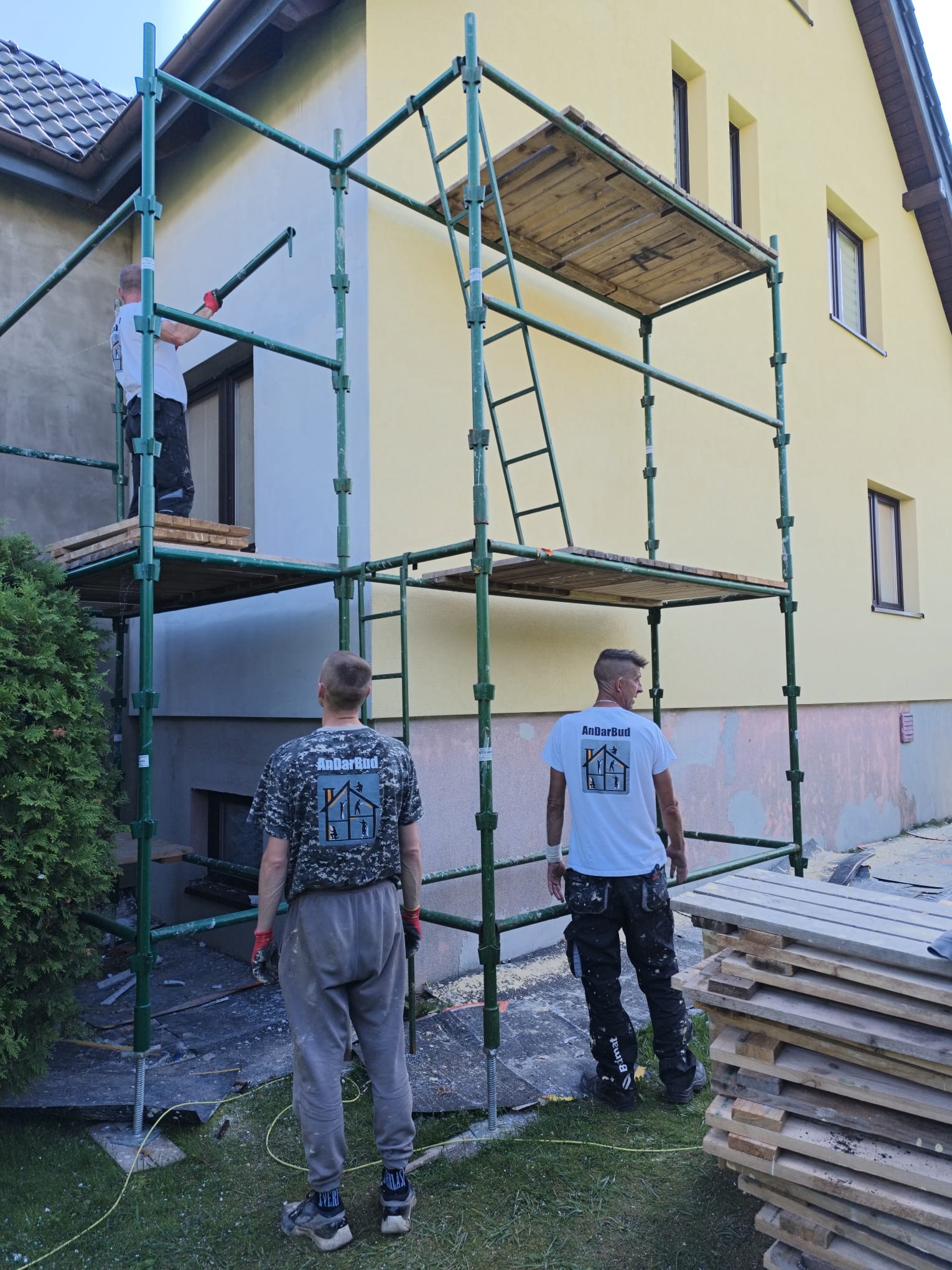 Żółta elewacja domu w trakcie prac. Widoczne rusztowania, pracownicy w koszulkach z logo firmy, przygotowane deski. Częściowo ocieplony fragment ściany.