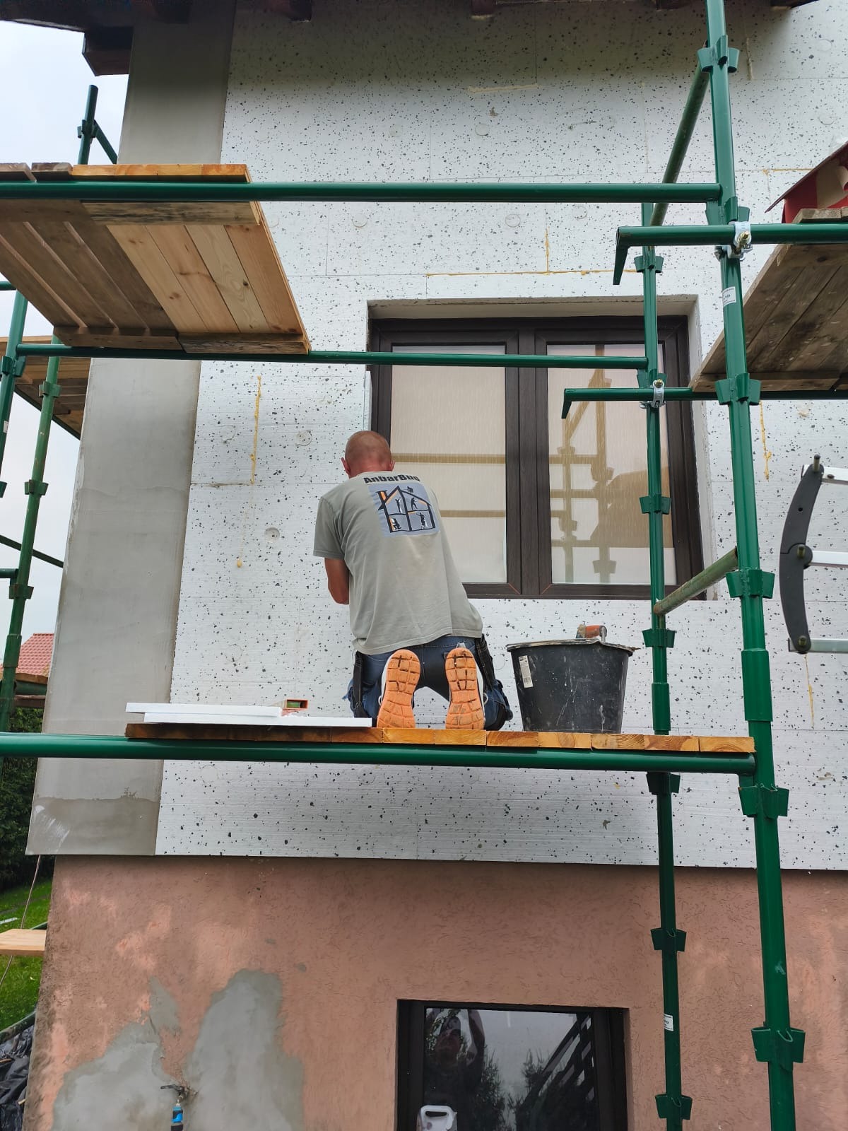 Pracownik na rusztowaniu montuje izolację termiczną na elewacji budynku. Widoczne płyty styropianowe i narzędzia. Ujęcie z dołu, skupione na detalu prac.