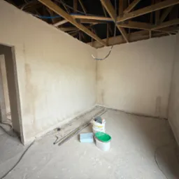 Wnętrze po tynkowaniu gipsowym: gładkie, jasne ściany, surowy strop z drewnianych belek, kable i puszki na podłodze, przygotowane do dalszych prac wykończeniowych.