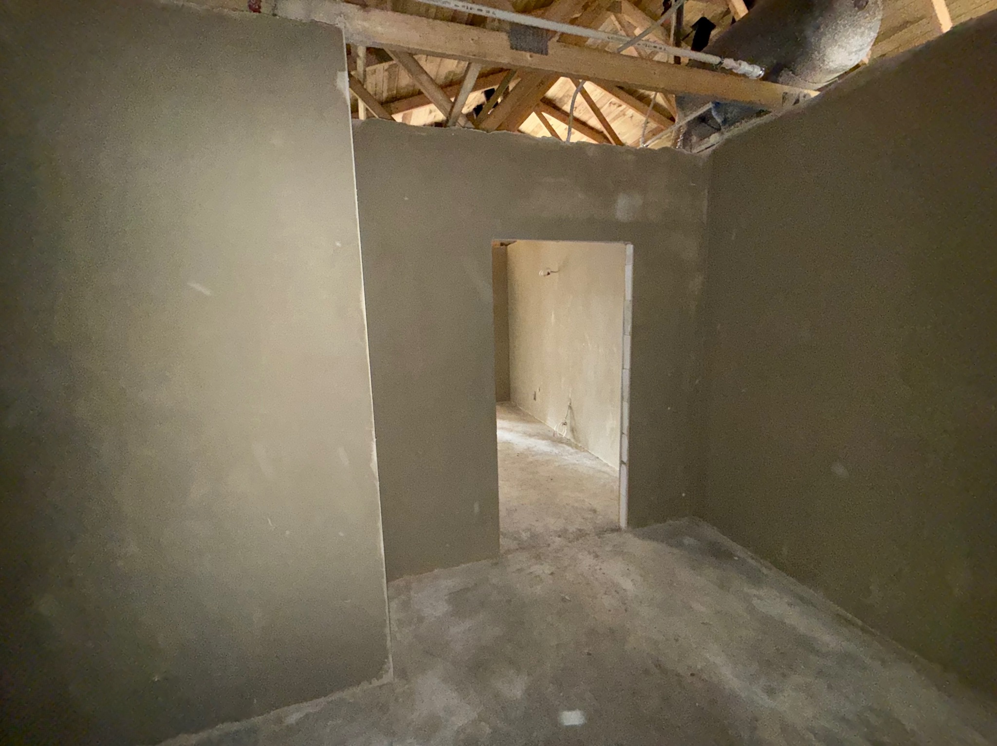 Wnętrze po tynkowaniu hybrydowym: gładkie, szare ściany, surowy strop drewniany, widoczne przejście do kolejnego pomieszczenia. Stan surowy po nałożeniu tynków.