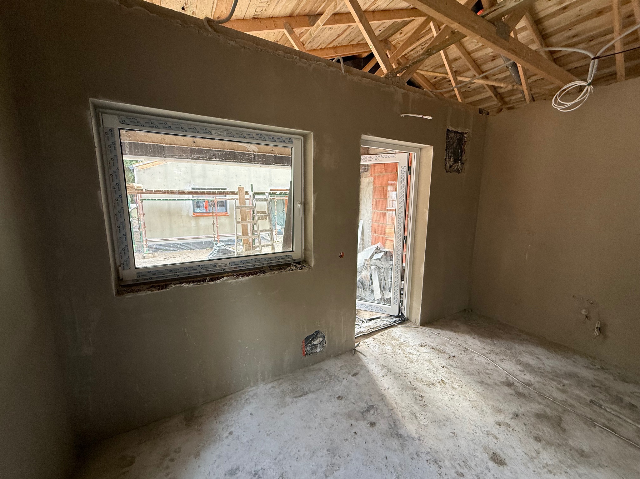 Wnętrze w trakcie tynkowania: ściany pokryte świeżym tynkiem, widoczne okno z rusztowaniem na zewnątrz, surowy strop drewniany i wystające kable elektryczne.
