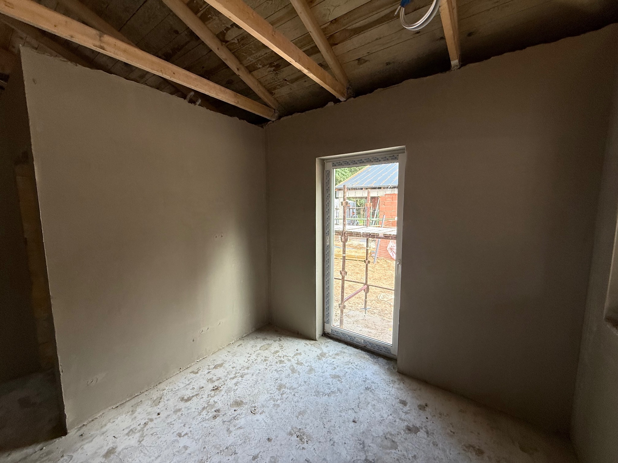 Pokój po tynkowaniu hybrydowym: gładkie, szare ściany, drewniany strop, okno z widokiem na rusztowanie. Widoczne niedoskonałości podłoża.