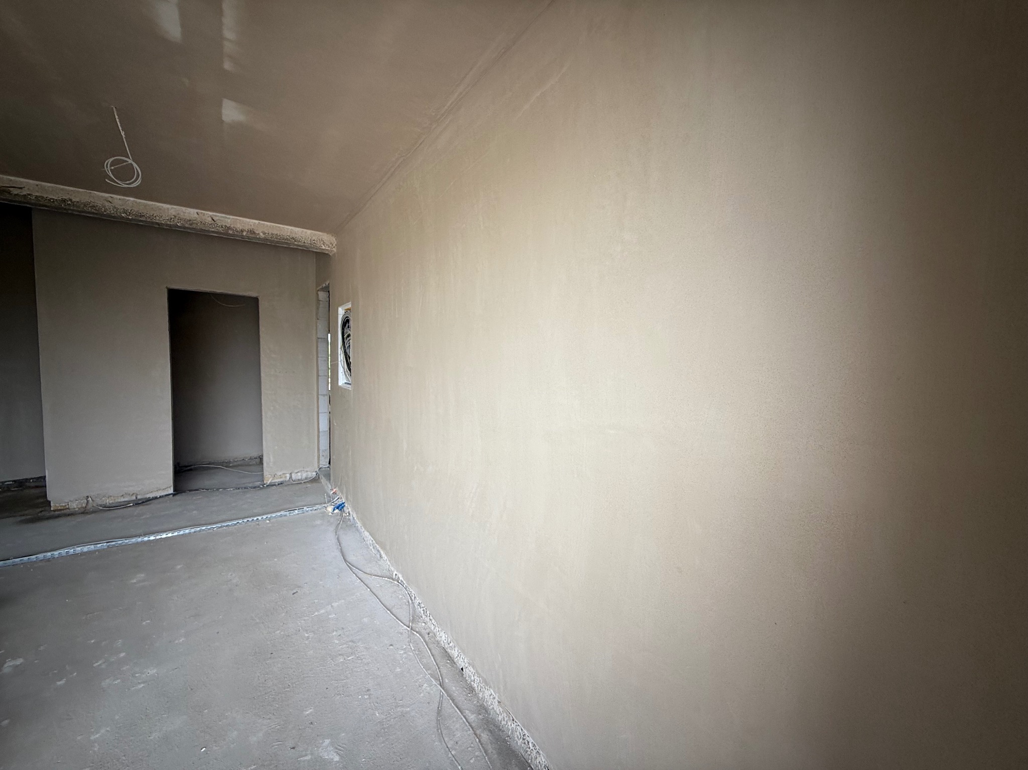 Wnętrze po tynkowaniu maszynowym: gładkie ściany, surowy beton na podłodze, widoczne przewody elektryczne, metalowe profile podłogowe.