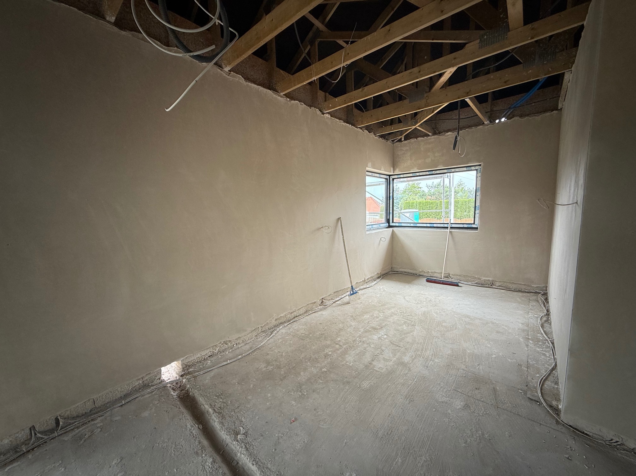 Wnętrze pokoju po nałożeniu tynków gipsowych na ściany. Widoczne kable elektryczne przy podłodze, drewniana konstrukcja dachu i okno z widokiem na zewnątrz.