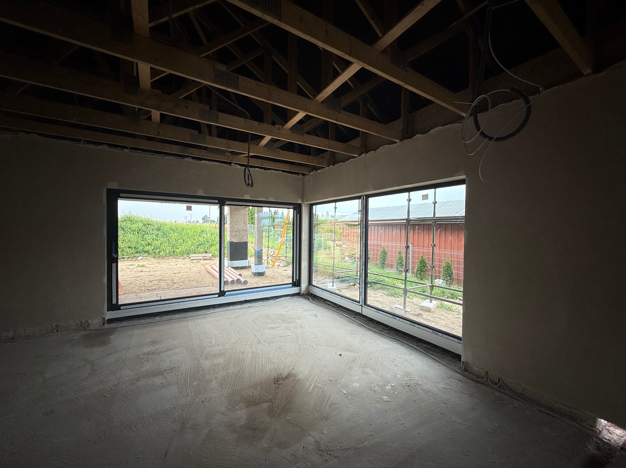 Jasne wnętrze po tynkowaniu gipsowym, z dużymi oknami przesuwnymi, widoczną konstrukcją dachu i surową podłogą. W tle zieleń.