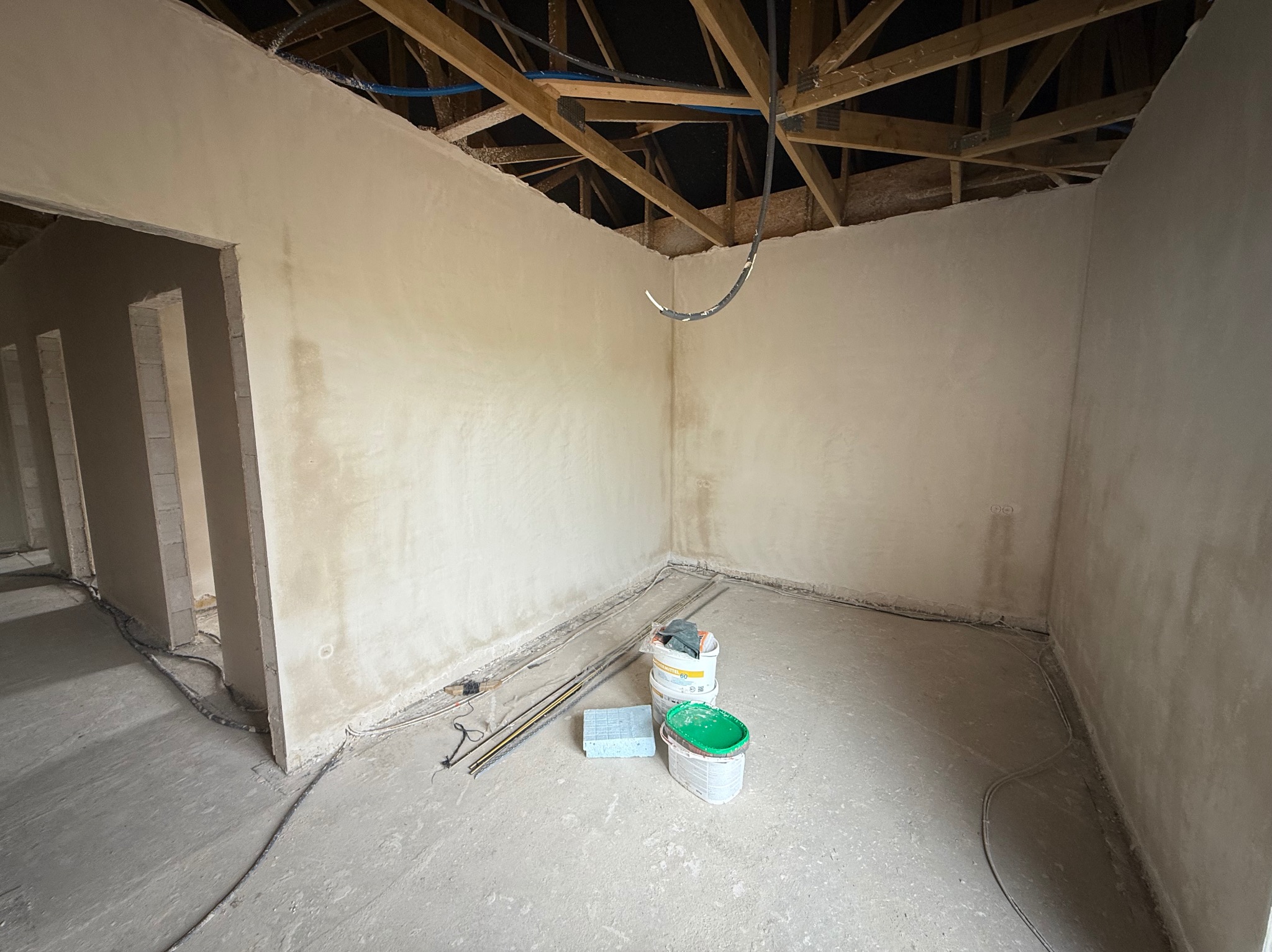 Wnętrze po tynkowaniu gipsowym: gładkie, jasne ściany, surowy strop z drewnianych belek, kable i puszki na podłodze, przygotowane do dalszych prac wykończeniowych.