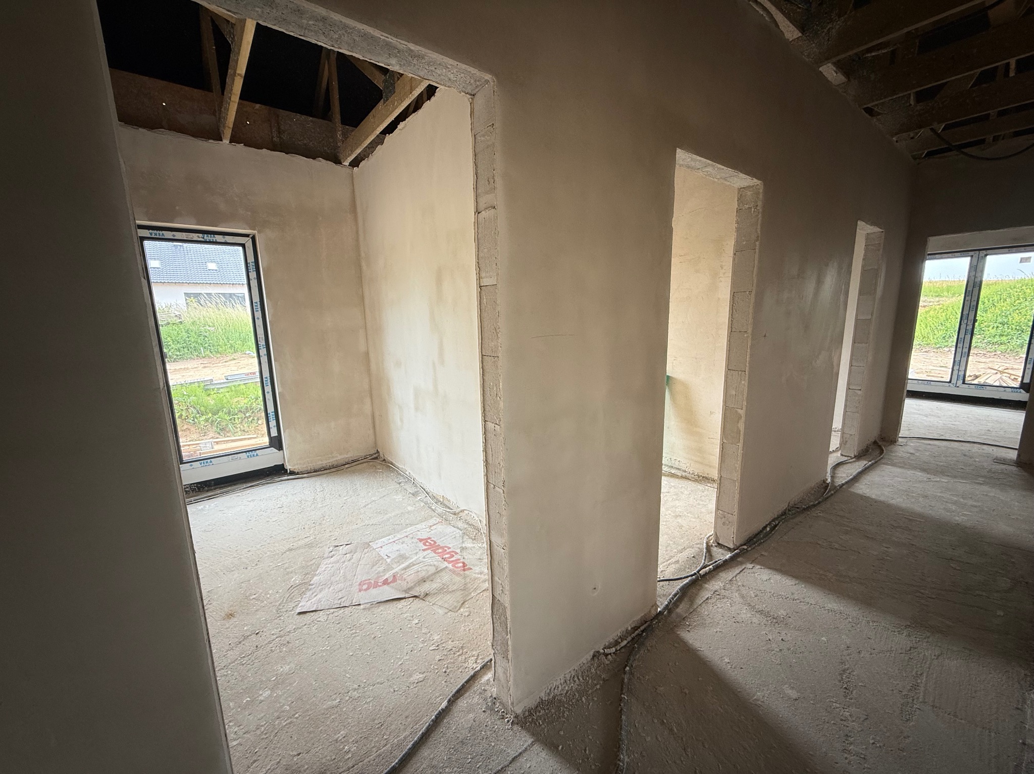 Wnętrze w trakcie tynkowania maszynowego. Widoczne surowe ściany pokryte świeżym tynkiem gipsowym, okna z widokiem na zieleń, odkryta konstrukcja dachu i przewody na podłodze.