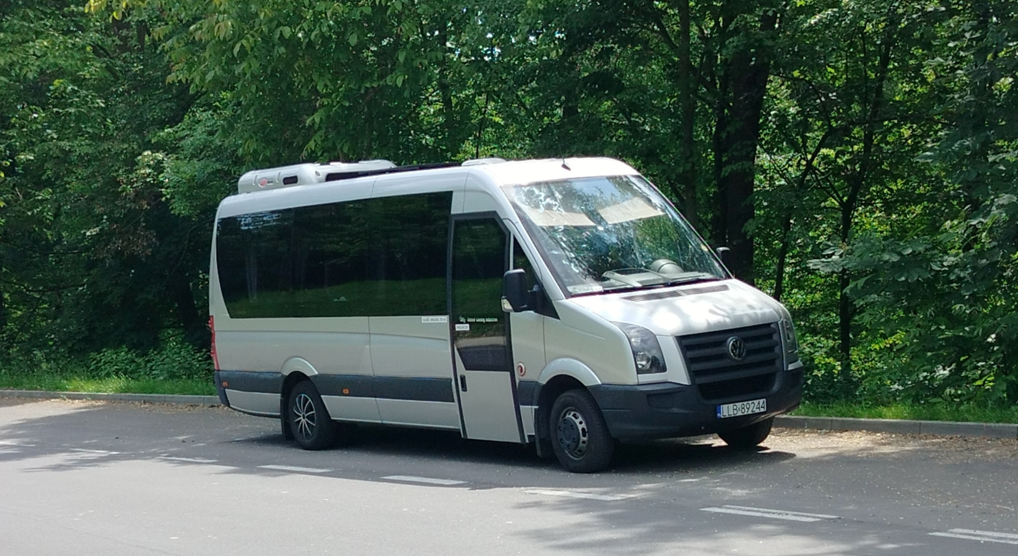 Srebrny bus z otwartymi drzwiami na tle zieleni. Widoczny numer rejestracyjny LLB 89244. Bus stoi na poboczu drogi.