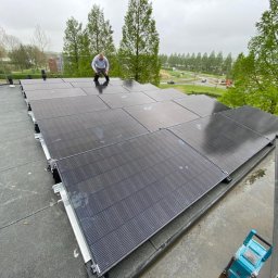 ELEKTRO - Dariusz - Montaż paneli fotowoltaicznych na płaskim dachu, widok z góry na instalatora pracującego na panelach, drabina oparta o krawędź dachu. Tło: zieleń i budynki.