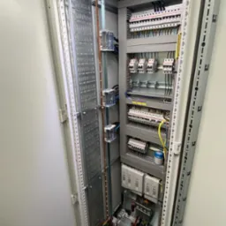 Otwarta szafa sterownicza z widocznymi elementami instalacji elektrycznej: bezpieczniki, przewody i szyny. Oświetlenie LED wewnątrz szafy. Widoczne narzędzia na podłodze.