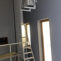 ELEKTRO - Dariusz - Biała klimatyzacja Mitsubishi na ścianie, widoczna drabina, dwa okna z żaluzjami, szare ściany i podłoga z ciemnych płytek. W tle stół roboczy.