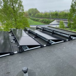 ELEKTRO - Dariusz - Montaż paneli fotowoltaicznych na płaskim dachu z widokiem na zieleń i infrastrukturę drogową w tle. Panele częściowo zamontowane na konstrukcji wsporczej.