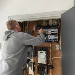 ELEKTRO - Dariusz - Elektryk w szarej bluzie montuje bezpieczniki w skrzynce elektrycznej na drewnianej ścianie z widocznymi przewodami. W tle biały sufit z kamerą.
