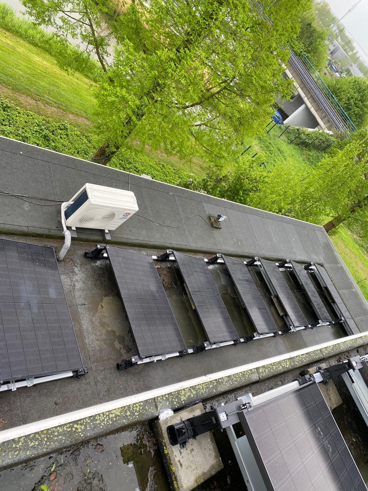 Instalacja fotowoltaiczna na dachu z klimatyzatorem, widok z góry na tle zieleni. Panele ustawione rzędami, klimatyzator biały, dach pokryty papą.