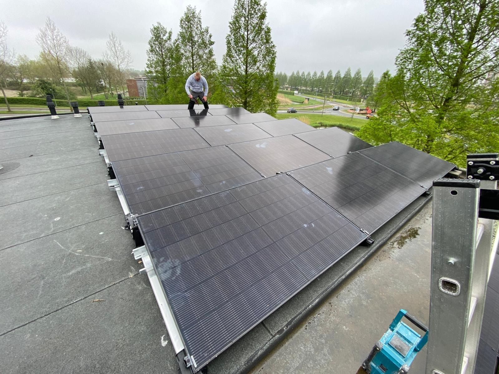 Montaż paneli fotowoltaicznych na płaskim dachu, widok z góry na instalatora pracującego na panelach, drabina oparta o krawędź dachu. Tło: zieleń i budynki.