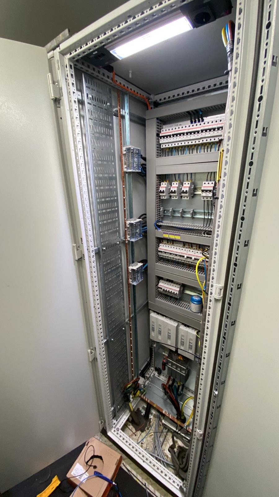 Otwarta szafa sterownicza z widocznymi elementami instalacji elektrycznej: bezpieczniki, przewody i szyny. Oświetlenie LED wewnątrz szafy. Widoczne narzędzia na podłodze.