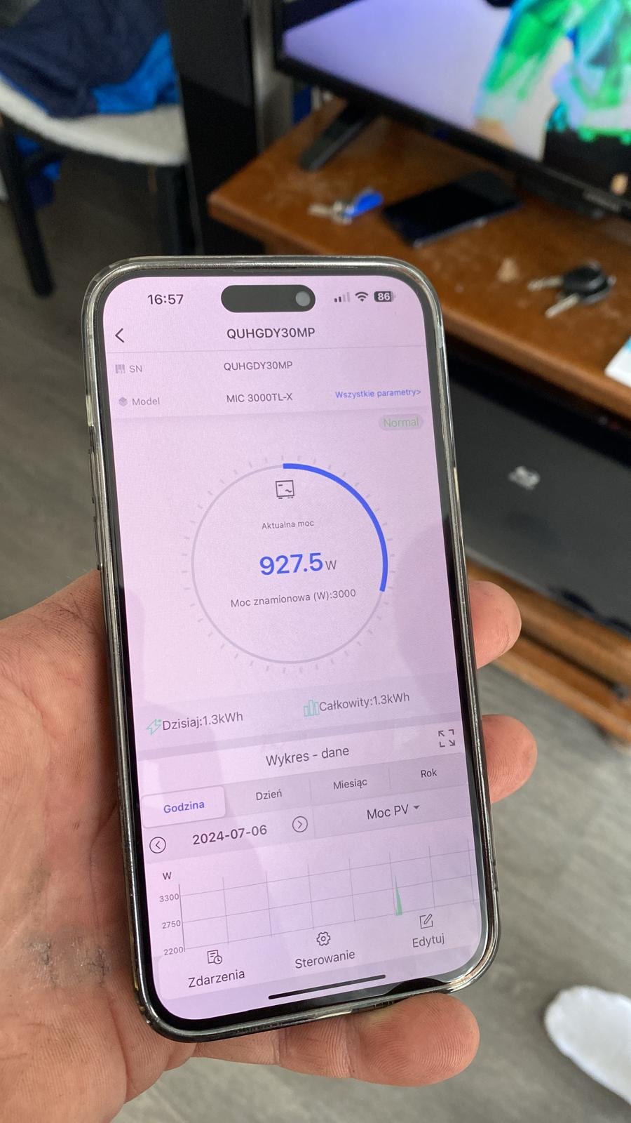 Telefon w dłoni prezentuje aplikację z wykresem mocy fotowoltaicznej. Wyświetlona aktualna moc 927.5W i data 2024-07-06.