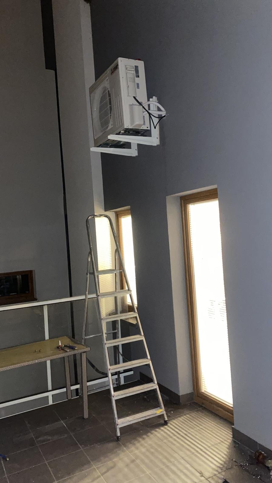 Biała klimatyzacja Mitsubishi na ścianie, widoczna drabina, dwa okna z żaluzjami, szare ściany i podłoga z ciemnych płytek. W tle stół roboczy.