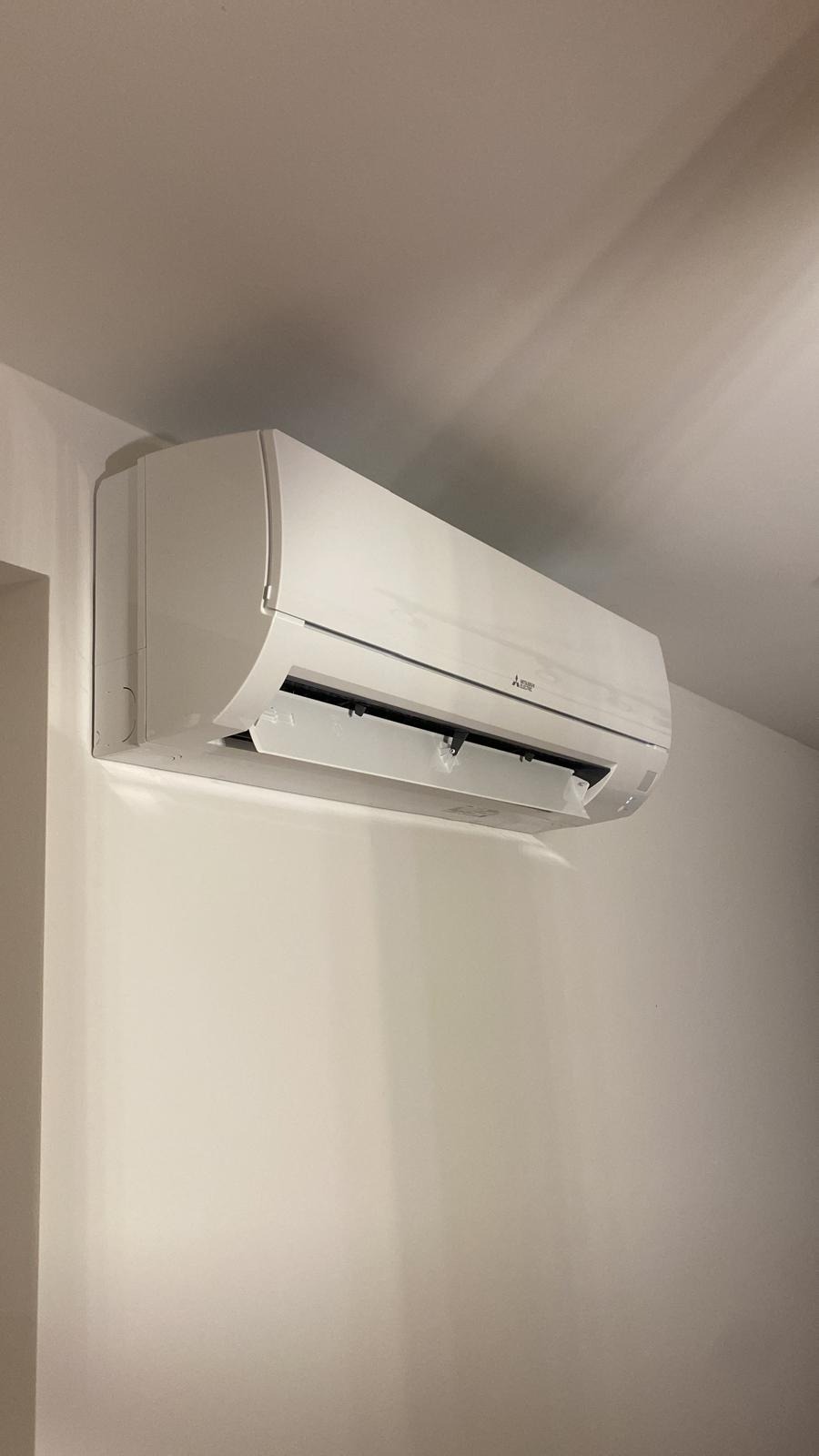 Biała klimatyzacja Mitsubishi zamontowana na ścianie, widoczna z boku, z otwartą klapką. Minimalistyczny design, jasne wnętrze, neutralne tło.