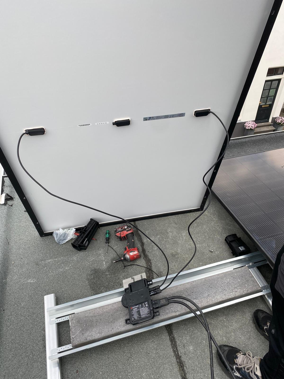 Montaż paneli fotowoltaicznych na dachu: widoczne panele, inwerter, narzędzia (wkrętarka) i fragment budynku w tle. Elementy instalacji w trakcie podłączania.