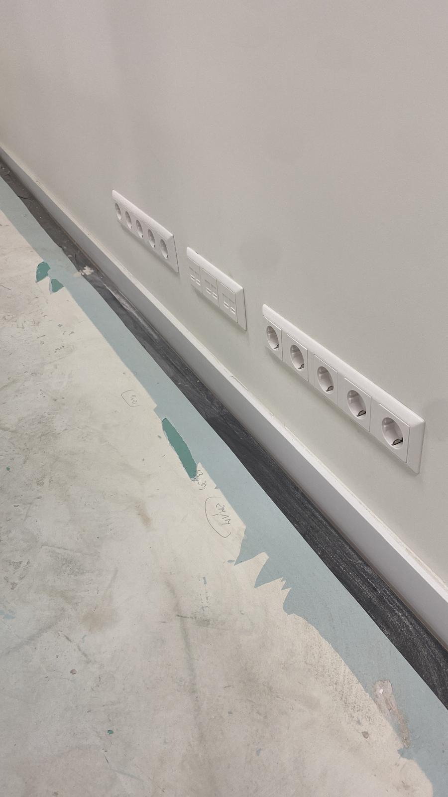 Białe gniazdka elektryczne i włączniki zamontowane w rzędzie na jasnej ścianie w pomieszczeniu w trakcie wykańczania. Widoczny fragment podłogi z resztkami folii ochronnej.