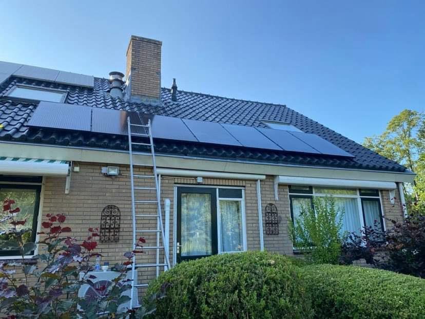 Dom jednorodzinny z panelami fotowoltaicznymi na dachu, drabina oparta o budynek, ceglana elewacja, bujna roślinność w ogrodzie.