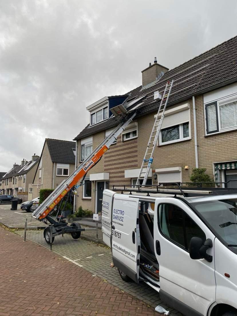 Montaż paneli słonecznych na dachu domu jednorodzinnego z użyciem podnośnika i drabiny, widoczny samochód dostawczy firmy instalacyjnej.