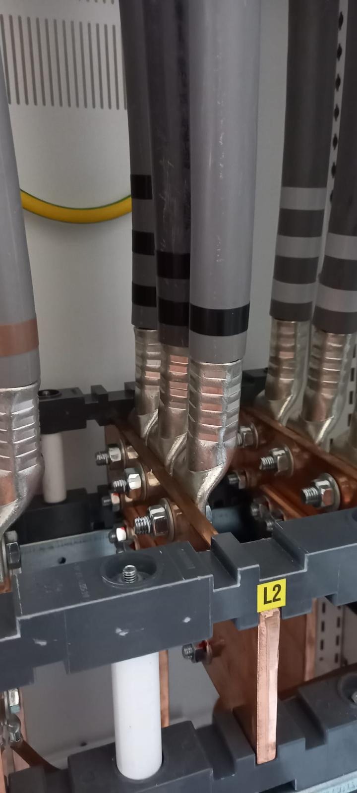 Szczegółowe ujęcie instalacji elektrycznej: przewody w szarych osłonach, miedziane złącza skręcane śrubami, etykieta 'L2'. Widok z bliska na elementy rozdzielni.