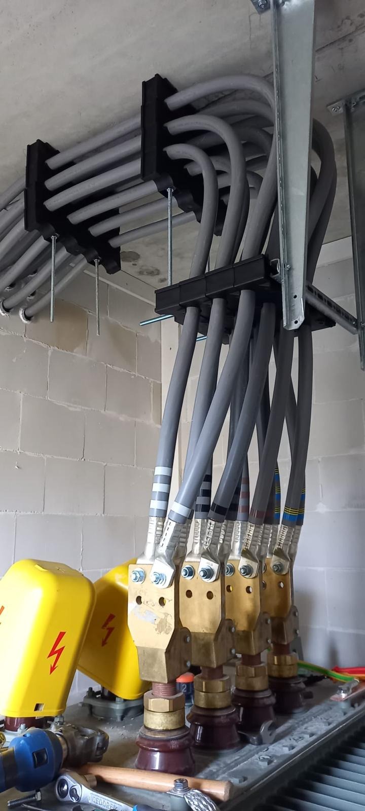 Szereg grubych kabli elektrycznych w szarych osłonach, przymocowanych do sufitu za pomocą czarnych uchwytów. Widoczne narzędzia i żółte elementy instalacji.