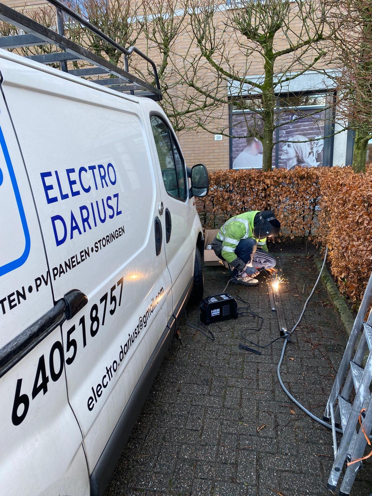 Pracownik w kombinezonie spawalniczym spawa metalową konstrukcję na zewnątrz, obok firmowego busa z napisem 'Electro Dariusz' i numerem telefonu.