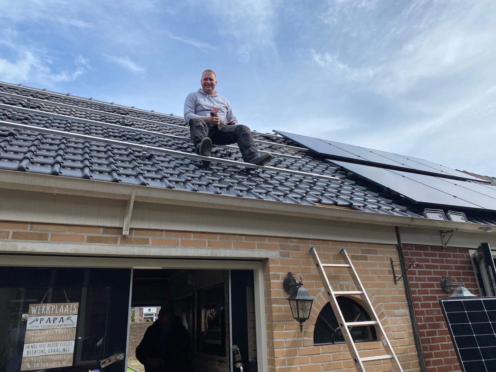 Instalator siedzi na dachu z panelami fotowoltaicznymi, trzymając narzędzia. Widoczna drabina oparta o budynek z cegły i częściowo zamontowane panele.