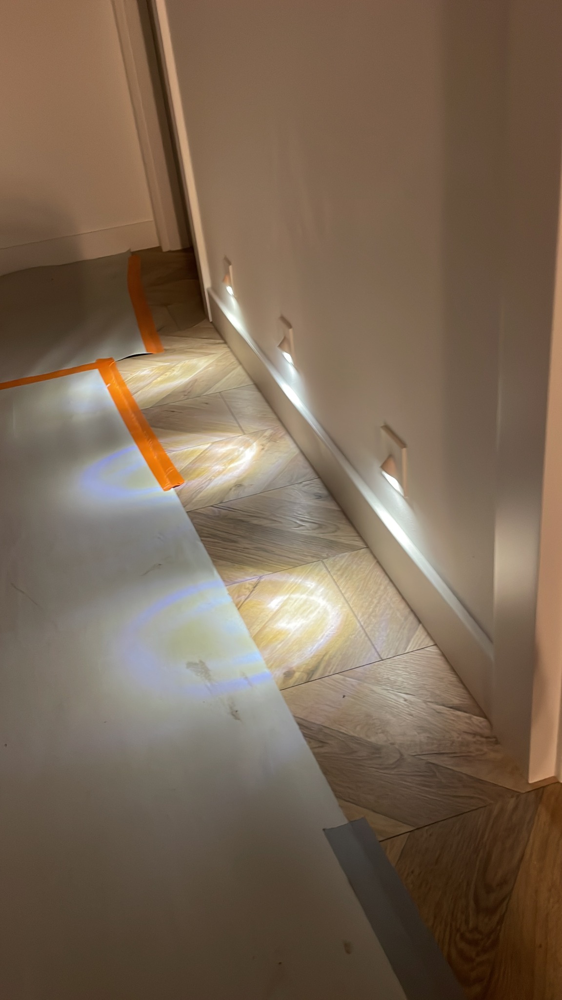 Podświetlane, kwadratowe lampy ścienne wzdłuż listwy przypodłogowej, rzucające światło na podłogę z wzorem jodełki, zabezpieczoną płytami z pomarańczową taśmą.