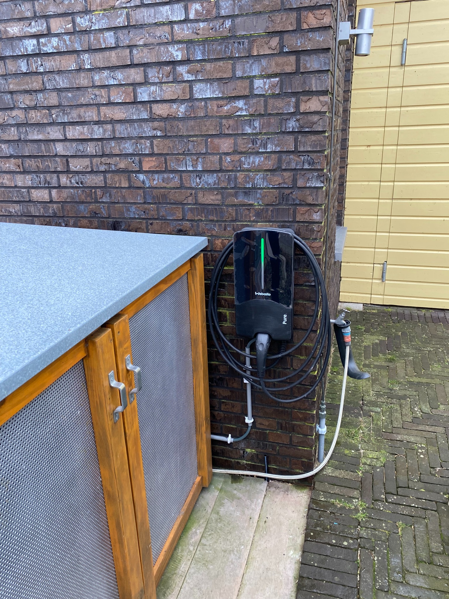 Czarna stacja ładowania Webasto Pure z zielonym paskiem LED, zamontowana na ceglanej ścianie zewnętrznej budynku, z podłączonym kablem. Obok drewniana szafka.