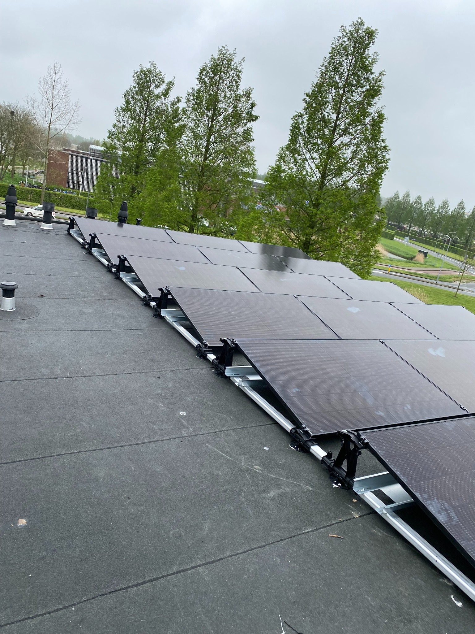Czarny dach z panelami fotowoltaicznymi w układzie rzędowym, wspartymi na srebrnych konstrukcjach. W tle zieleń drzew i budynek. Pochmurny dzień.
