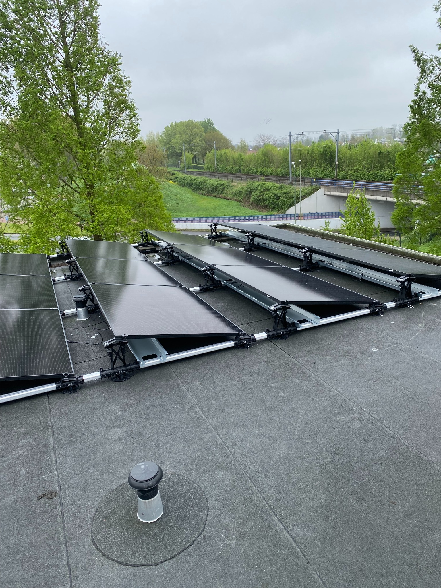 Montaż paneli fotowoltaicznych na płaskim dachu z widokiem na zieleń i infrastrukturę drogową w tle. Panele częściowo zamontowane na konstrukcji wsporczej.