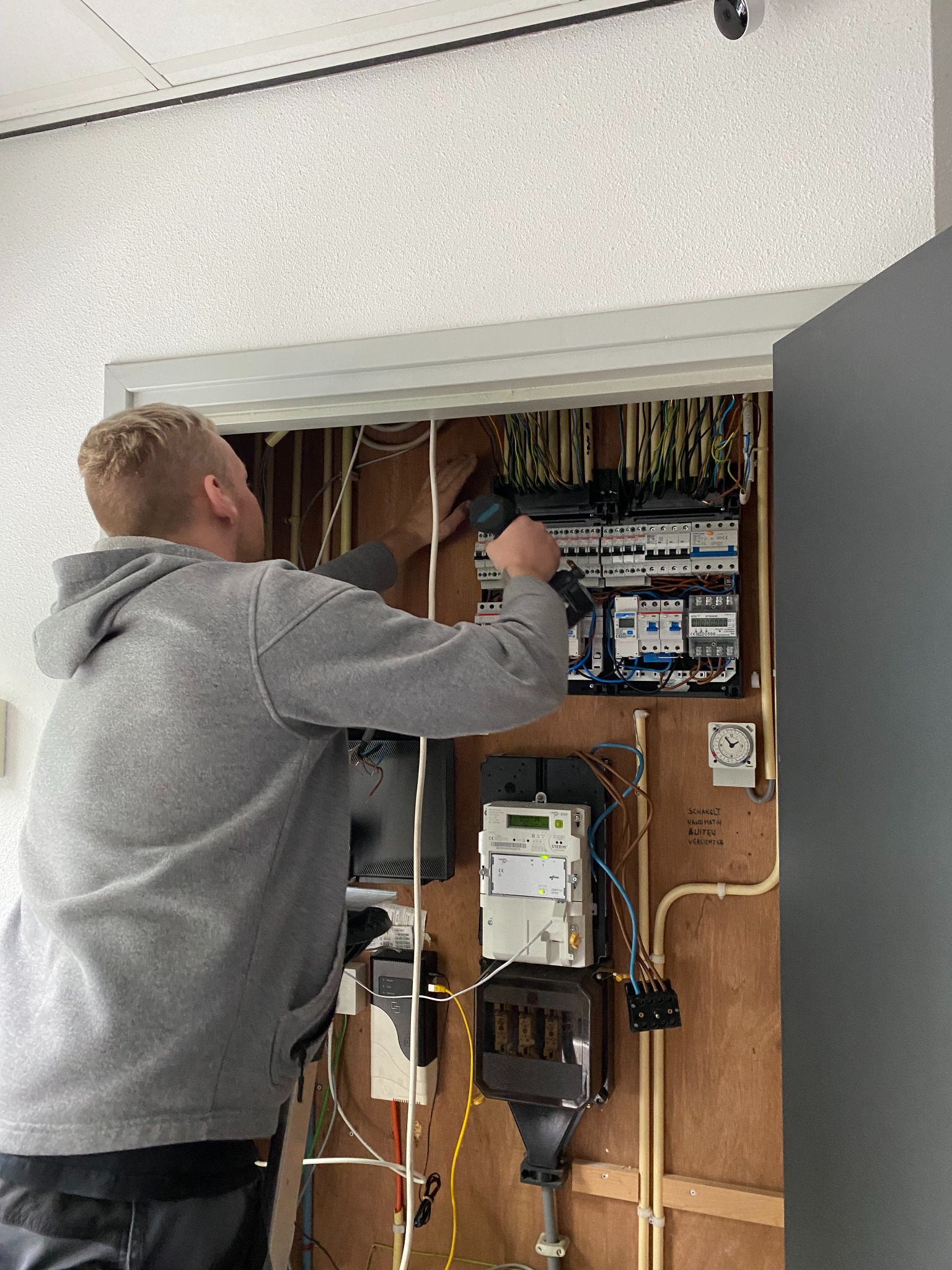 Elektryk w szarej bluzie montuje bezpieczniki w skrzynce elektrycznej na drewnianej ścianie z widocznymi przewodami. W tle biały sufit z kamerą.