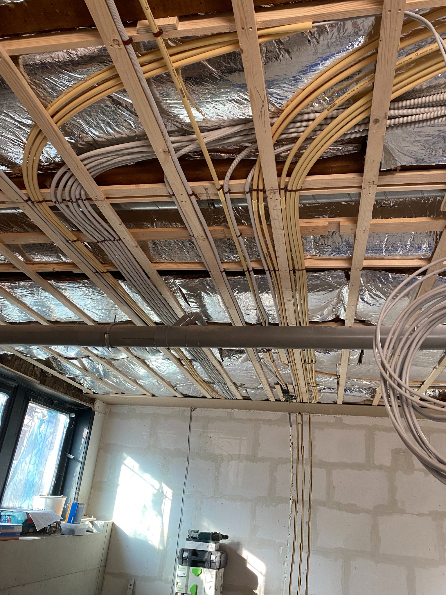 Instalacja elektryczna i rury wentylacyjne pod sufitem, widoczne kable w peszlach, drewniana konstrukcja, izolacja folią aluminiową, surowy stan wykończenia.